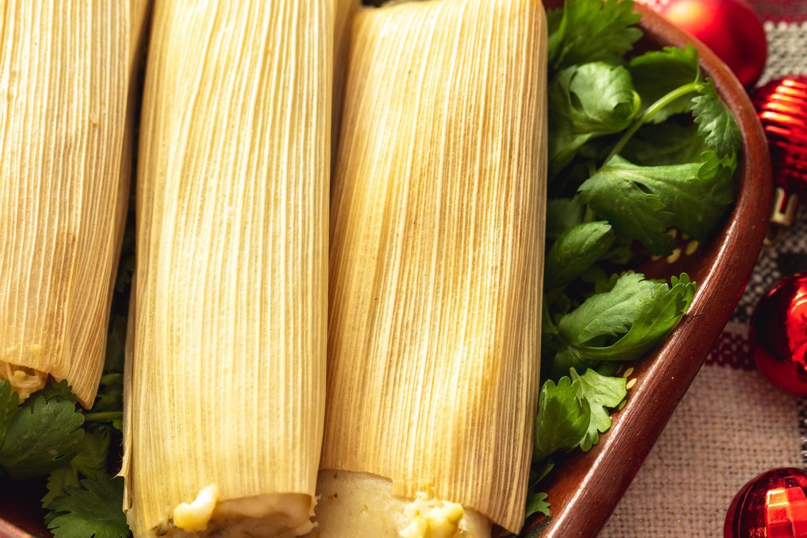 Golden tamales in corn husks on a folded cloth with cilantro and red chiles
